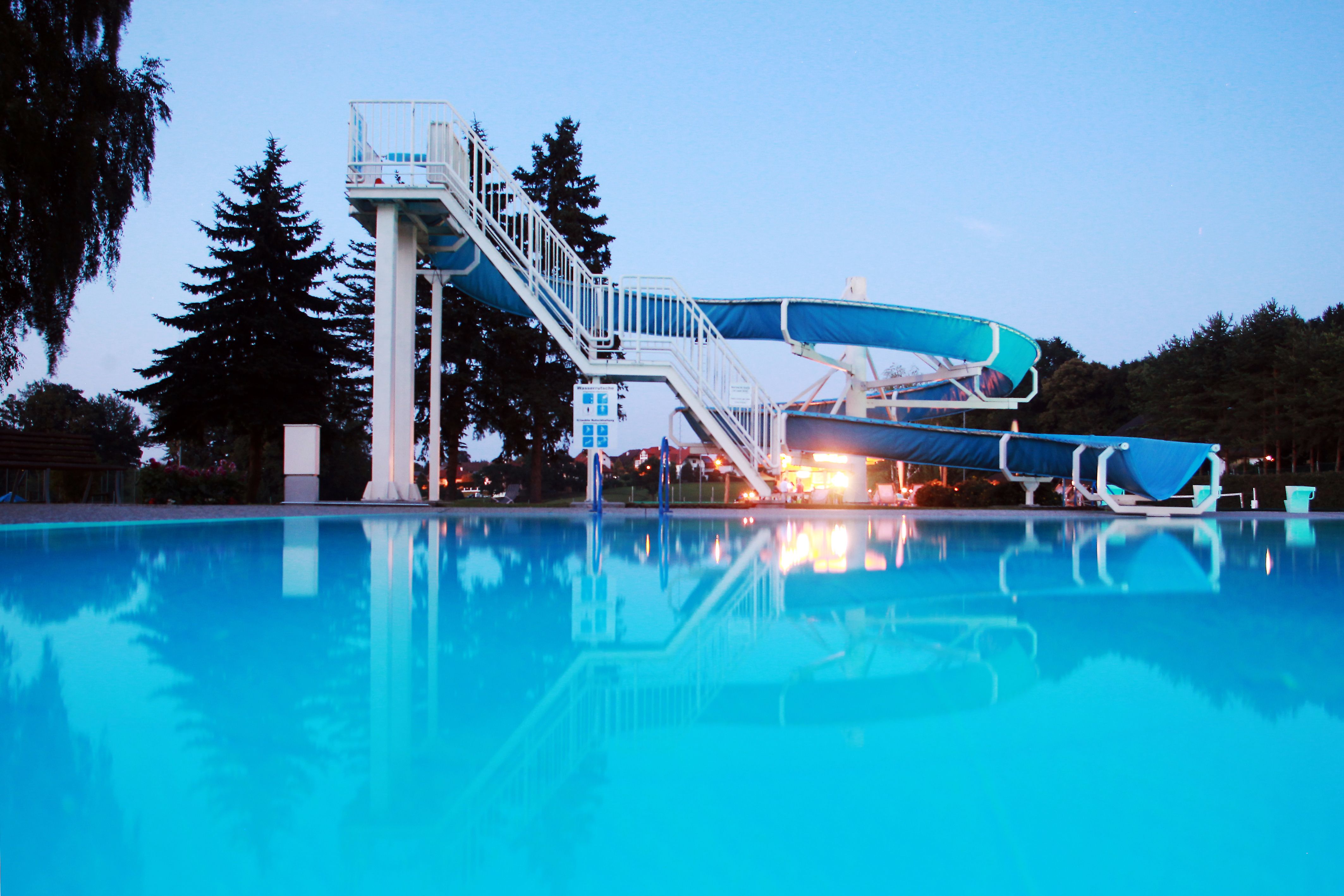 An outdoor pool with a large blue water slide and a clear pool in the foreground, surrounded by trees at dusk.