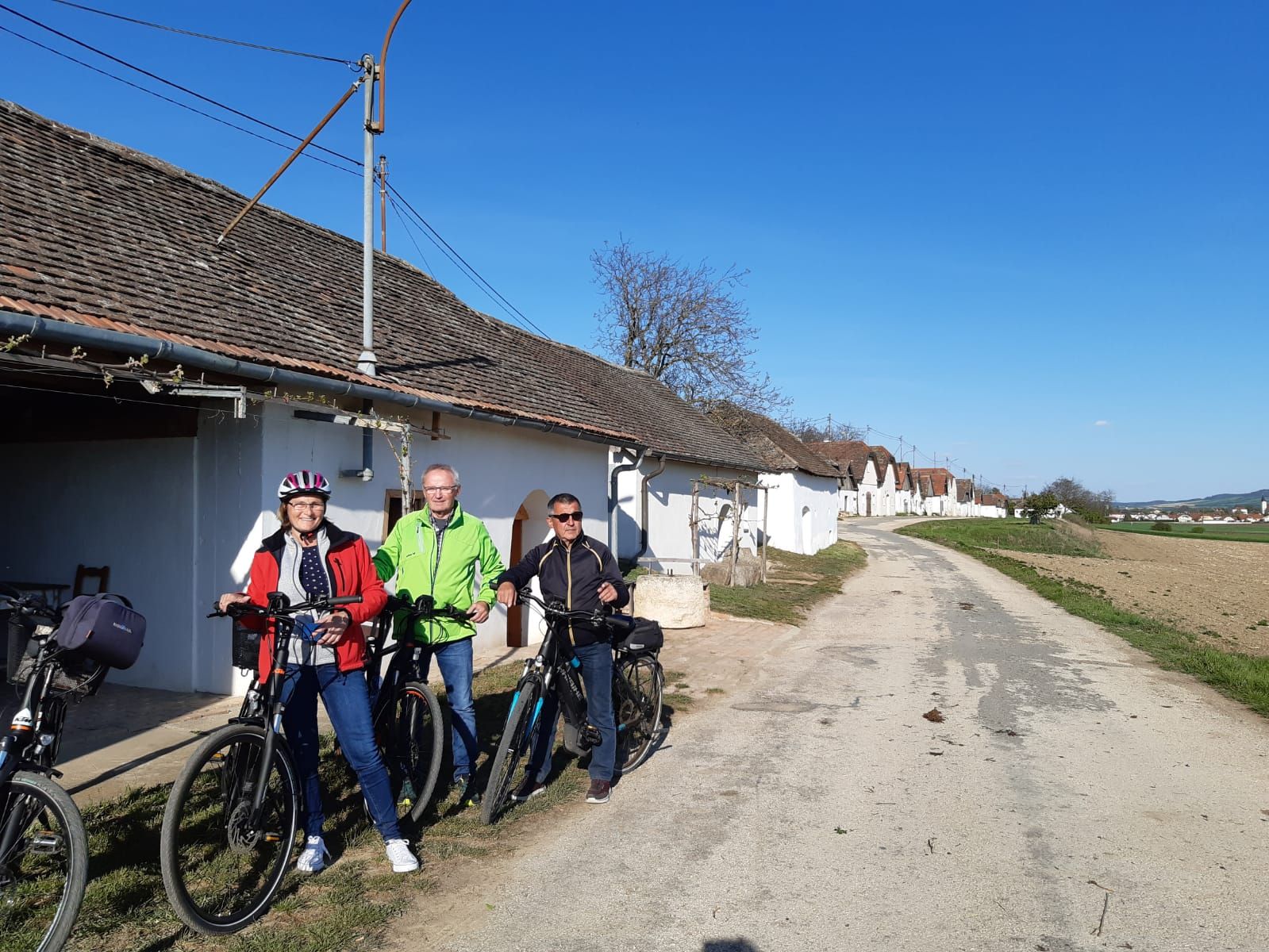 Three people with bicycles in front of a row of wine cellars in a rural setting.