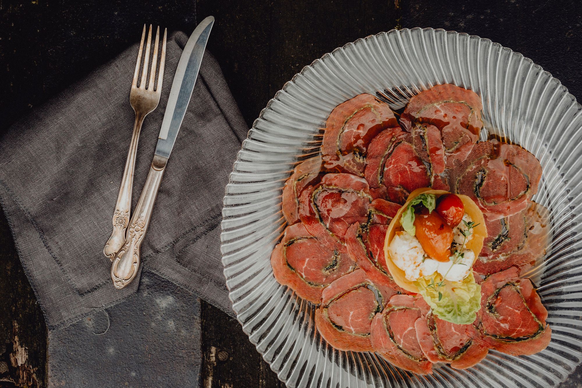 A plate with thinly sliced meat and a small salad garnish in the middle, cutlery on a napkin next to it.