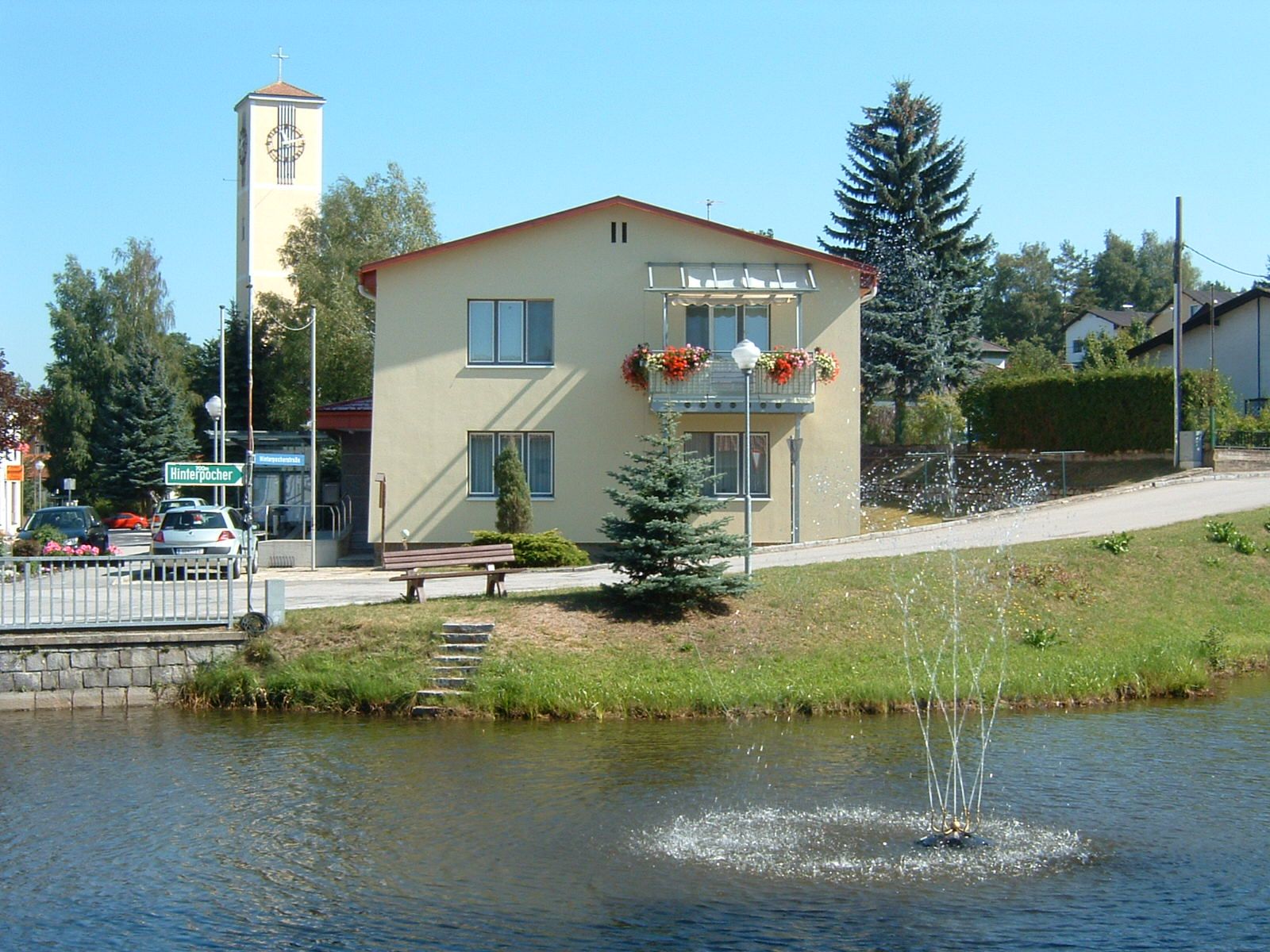 A yellow building with flower boxes and a bell tower in the background, with a pond and fountain in front of it.