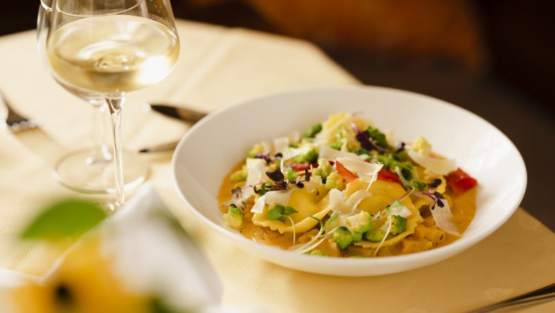 A plate of cream cheese ravioli, garnished with vegetables and herbs, next to a glass of white wine on a laid table.