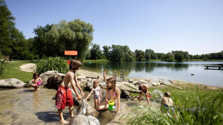 Children play in the shallow water of a lake with stones and plants in the background.