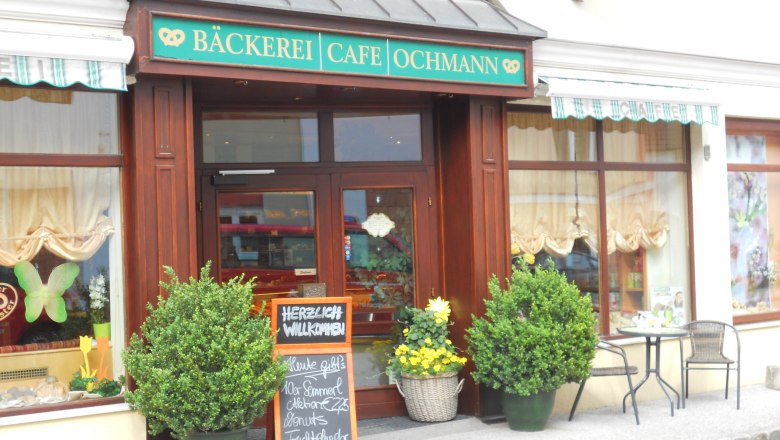 Entrance of a bakery, decorated with plants, a sign and a small table with chairs.