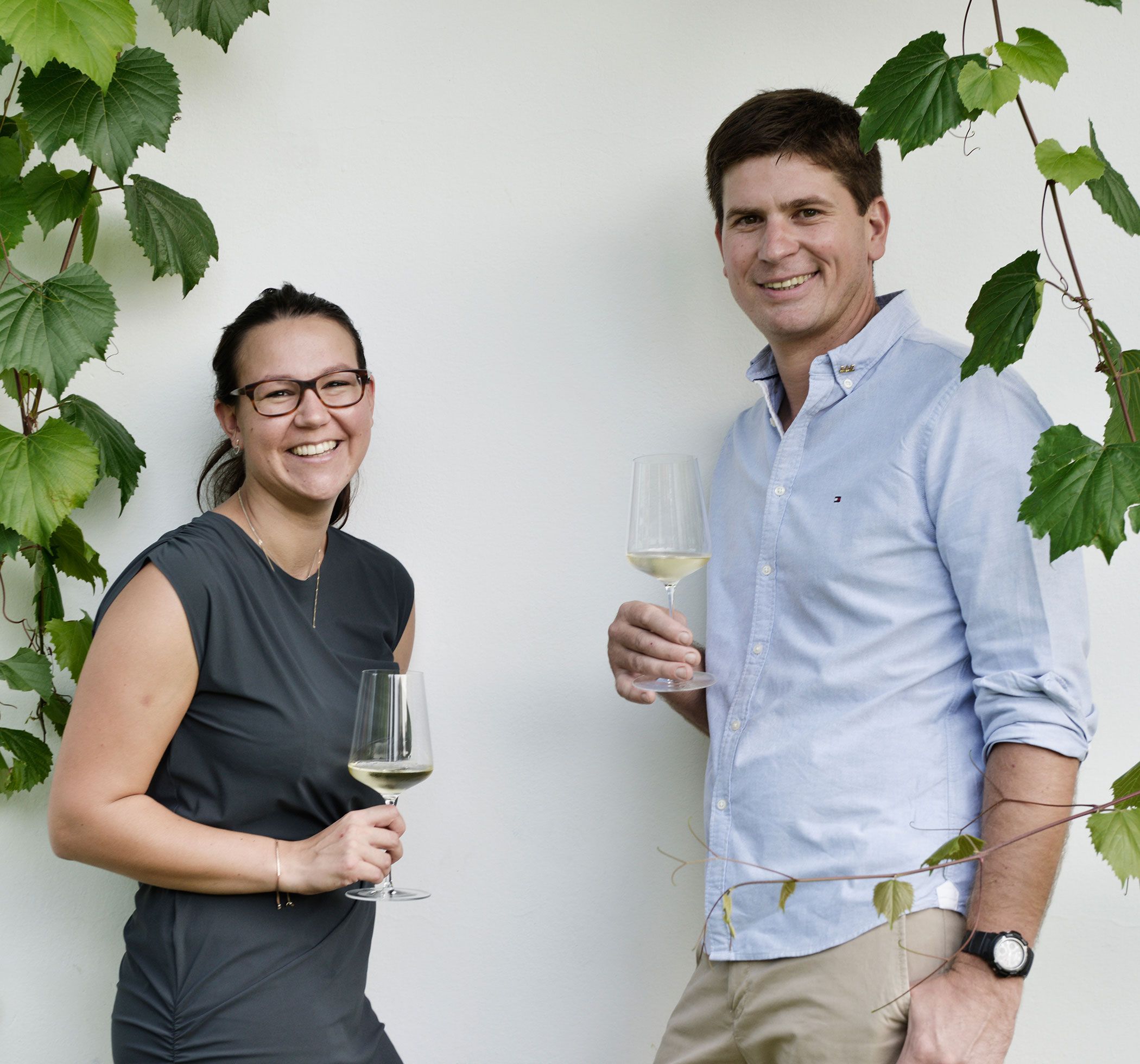 Two people with wine glasses in front of a white wall and grapevines.