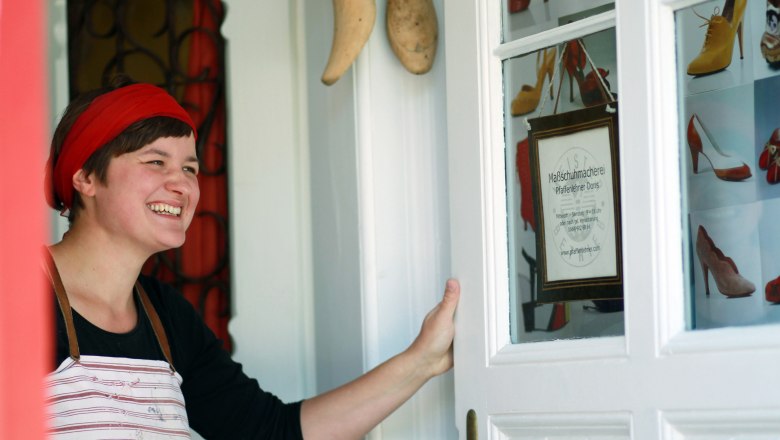 A woman with a red headband and apron stands smiling at a door with shoe pictures and a sign for the Pfaffenlehner Doris bespoke shoemaker's shop.