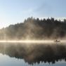 A boat on a misty lake in front of a wooded shore at sunrise.