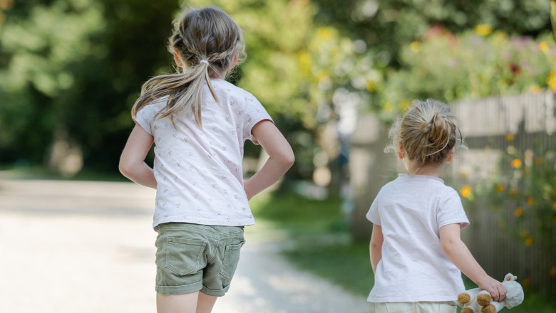 Two children run along a gravel path in a park, surrounded by trees and flowers.