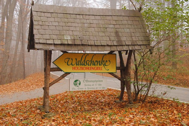 A wooden sign in the forest points to the Waldschenke Holzkohlengrill.