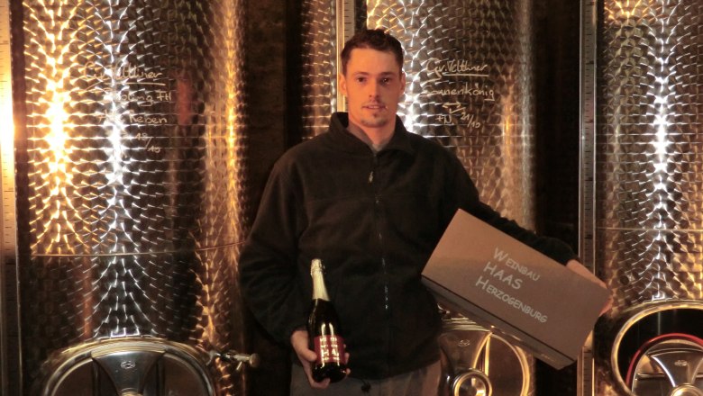A man stands in front of large stainless steel tanks holding a bottle of sparkling wine and a crate labeled 'Weinbau Haas Herzogenburg'.
