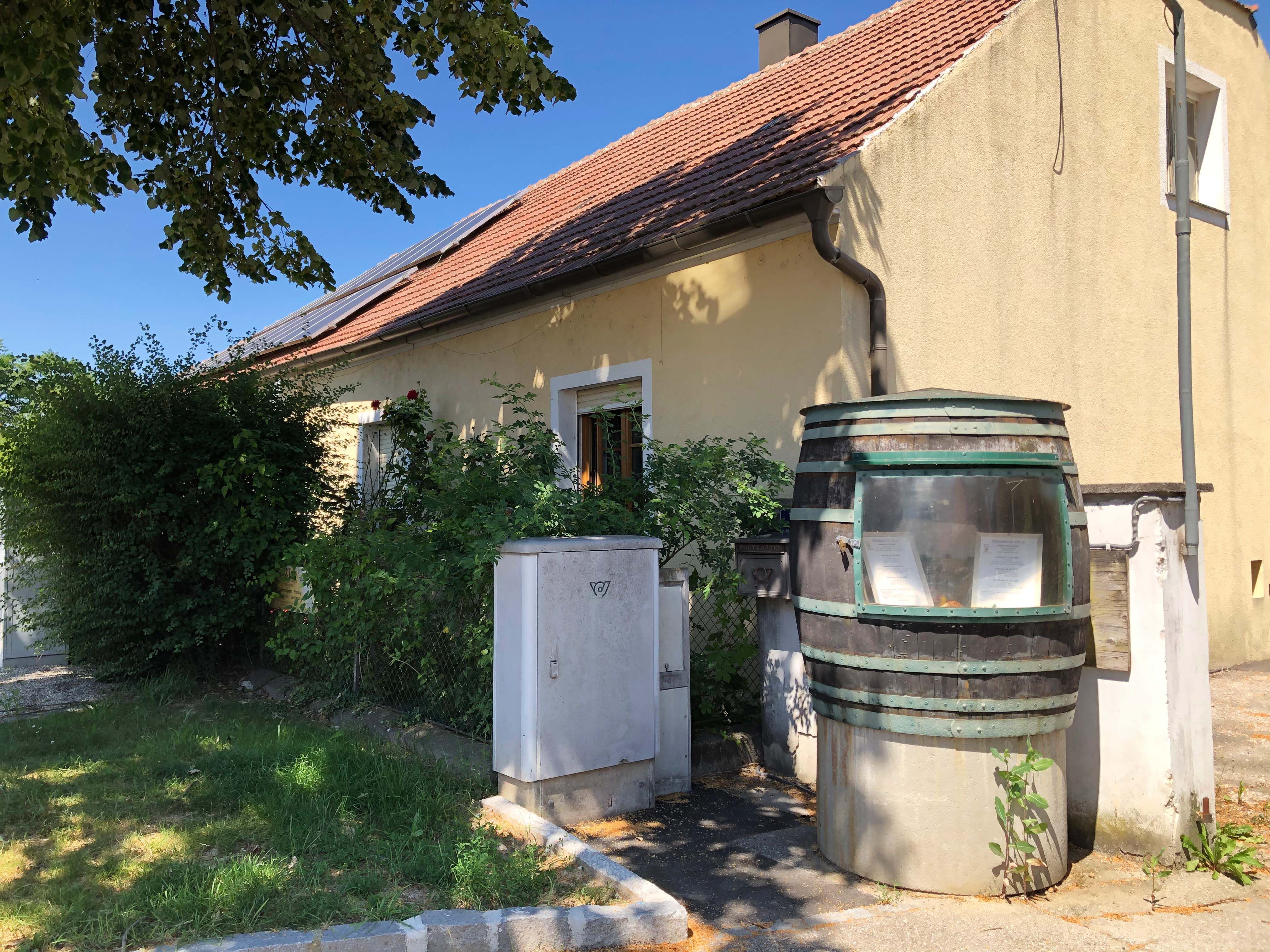 A yellow house with a red tiled roof and solar panels. In the foreground, a large barrel with a sight glass and plants.
