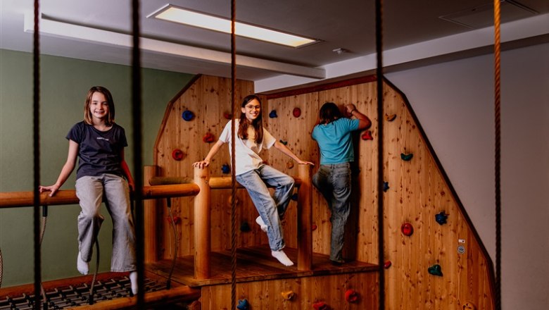 Children play in an indoor climbing area with wooden walls and colorful climbing holds.