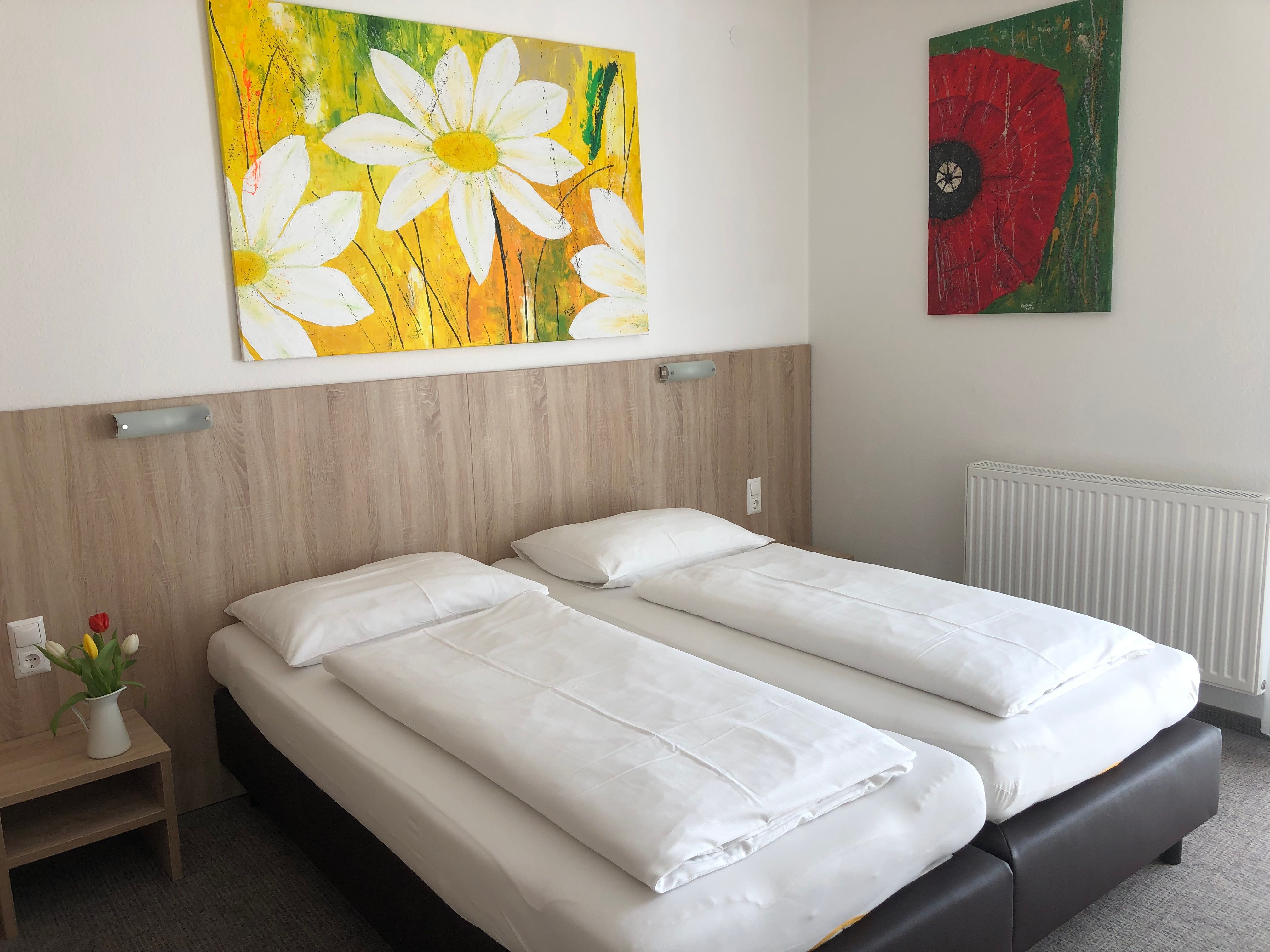 A simple hotel room with two single beds, a bedside table with flowers and two colorful paintings on the wall.
