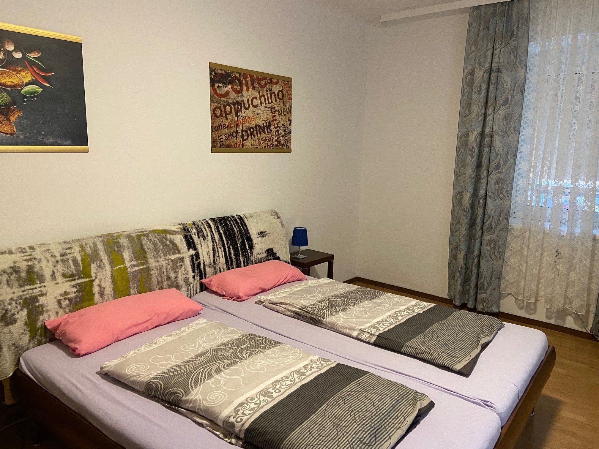 Bedroom with double bed, two pillows, two blankets, wall pictures and window with curtains.