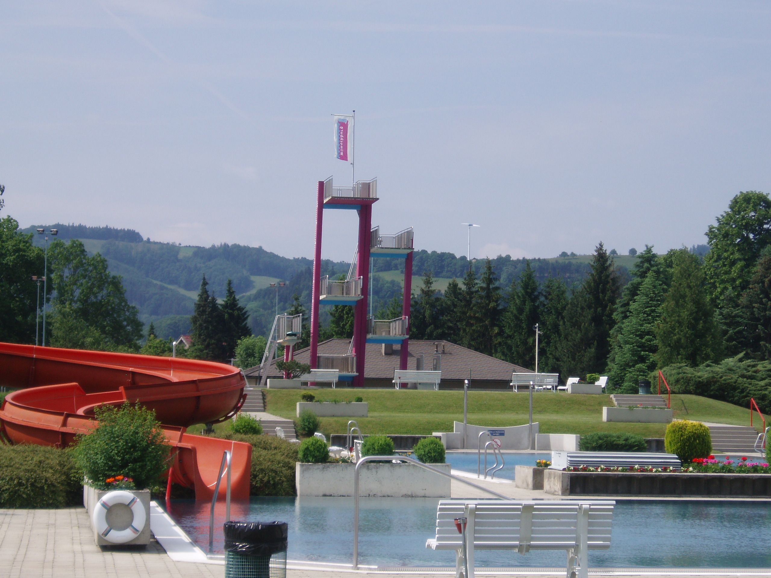 An outdoor pool with a slide, diving platform and green surroundings.