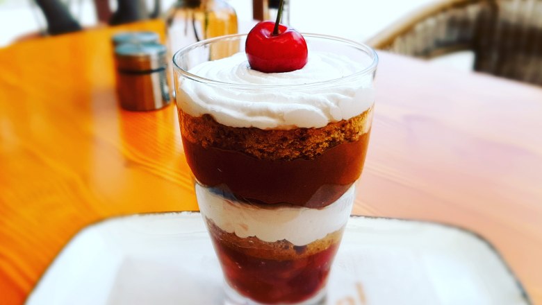 A dessert in a glass with layers of cream, cake and cherries, garnished with a cherry.