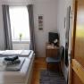 Bright bedroom with double bed, gray curtains, small desk and chair, TV and carpet on wooden floor.