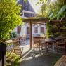 A sunny garden with wooden furniture and a covered seating area, surrounded by plants and a building in the background.