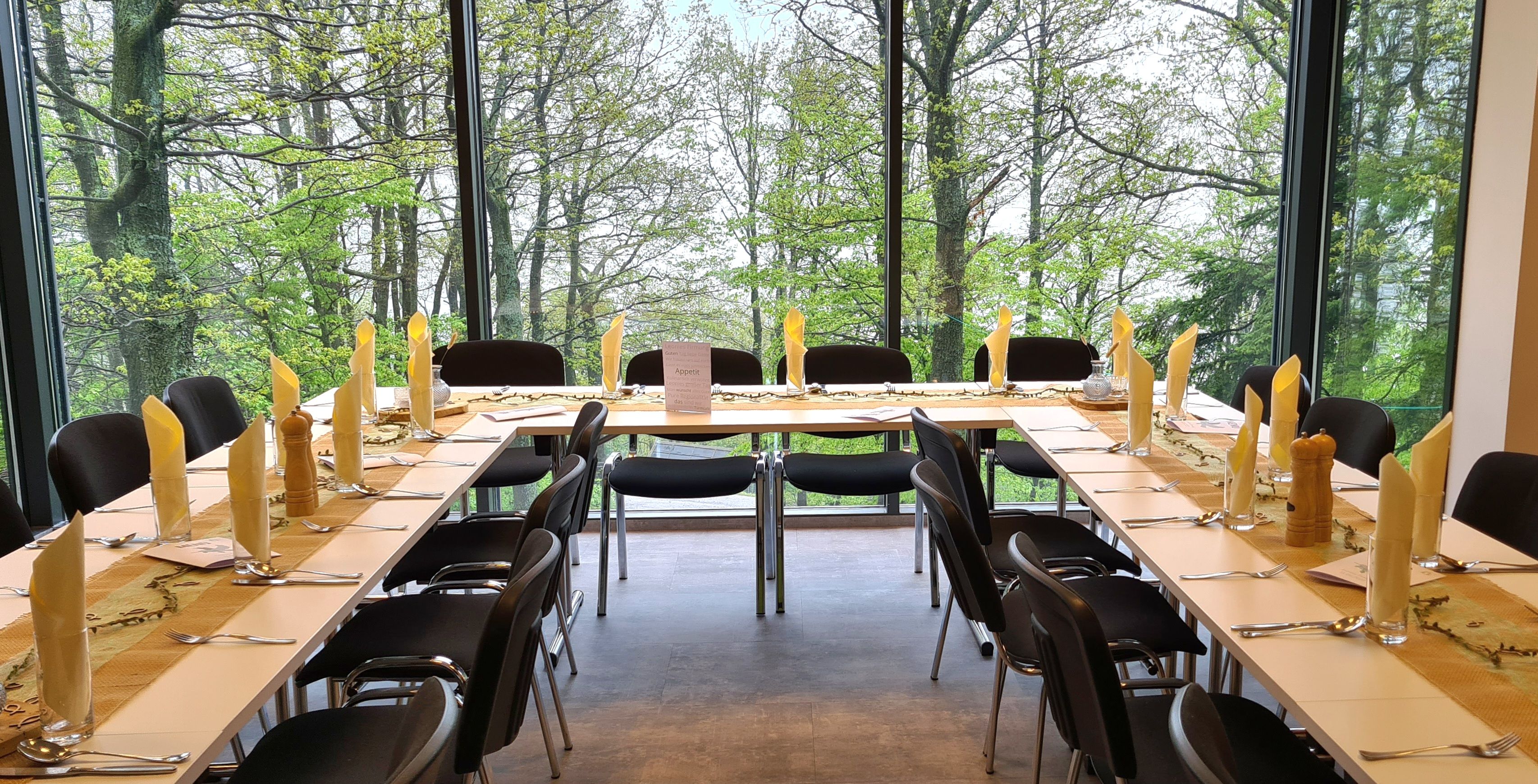 Set tables with yellow decorations and black chairs in front of a large window with a view of trees.