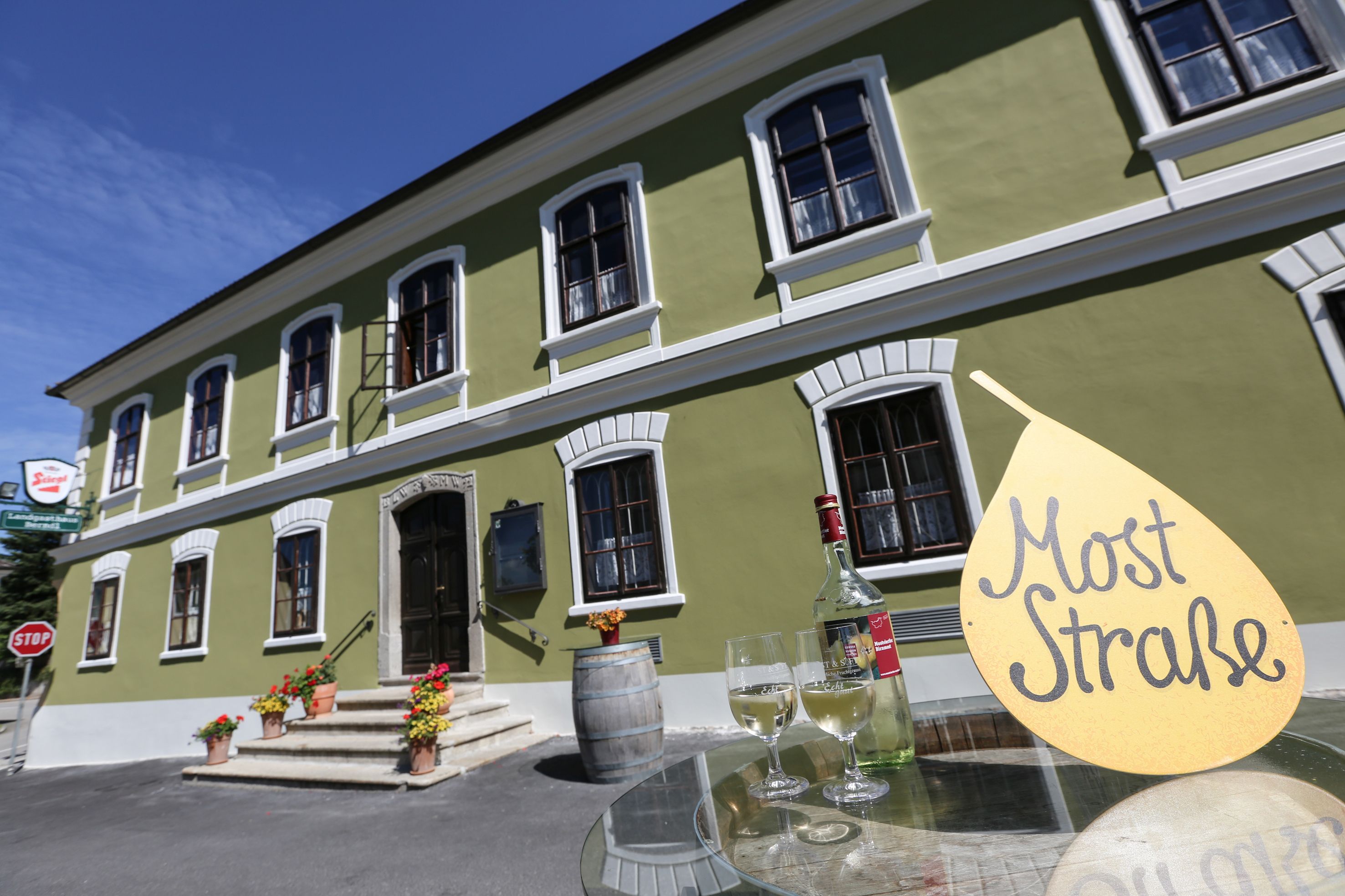 Green building with white windows, in front of it a table with a bottle of wine and glasses, sign with the inscription 'Cider Straße'.
