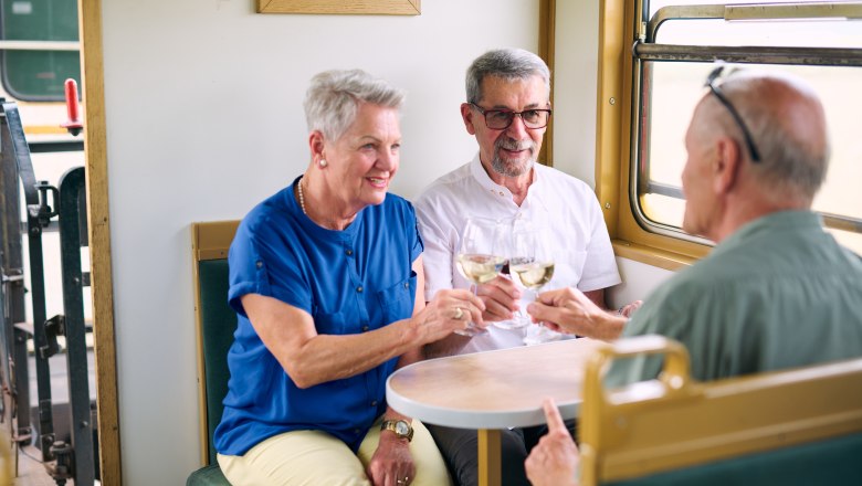 Three elderly people clink glasses of wine in a train compartment.