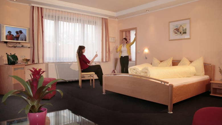 Two women in a hotel room with a double bed, one sitting, the other standing with her glass raised.