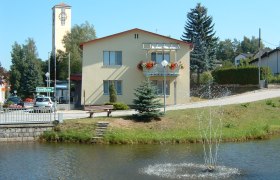 A yellow building with flower boxes and a bell tower in the background, with a pond and fountain in front of it.