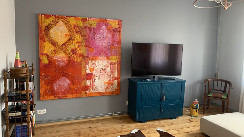 Living room with large colorful painting, TV on blue cabinet, wooden floor and cowhide carpet.
