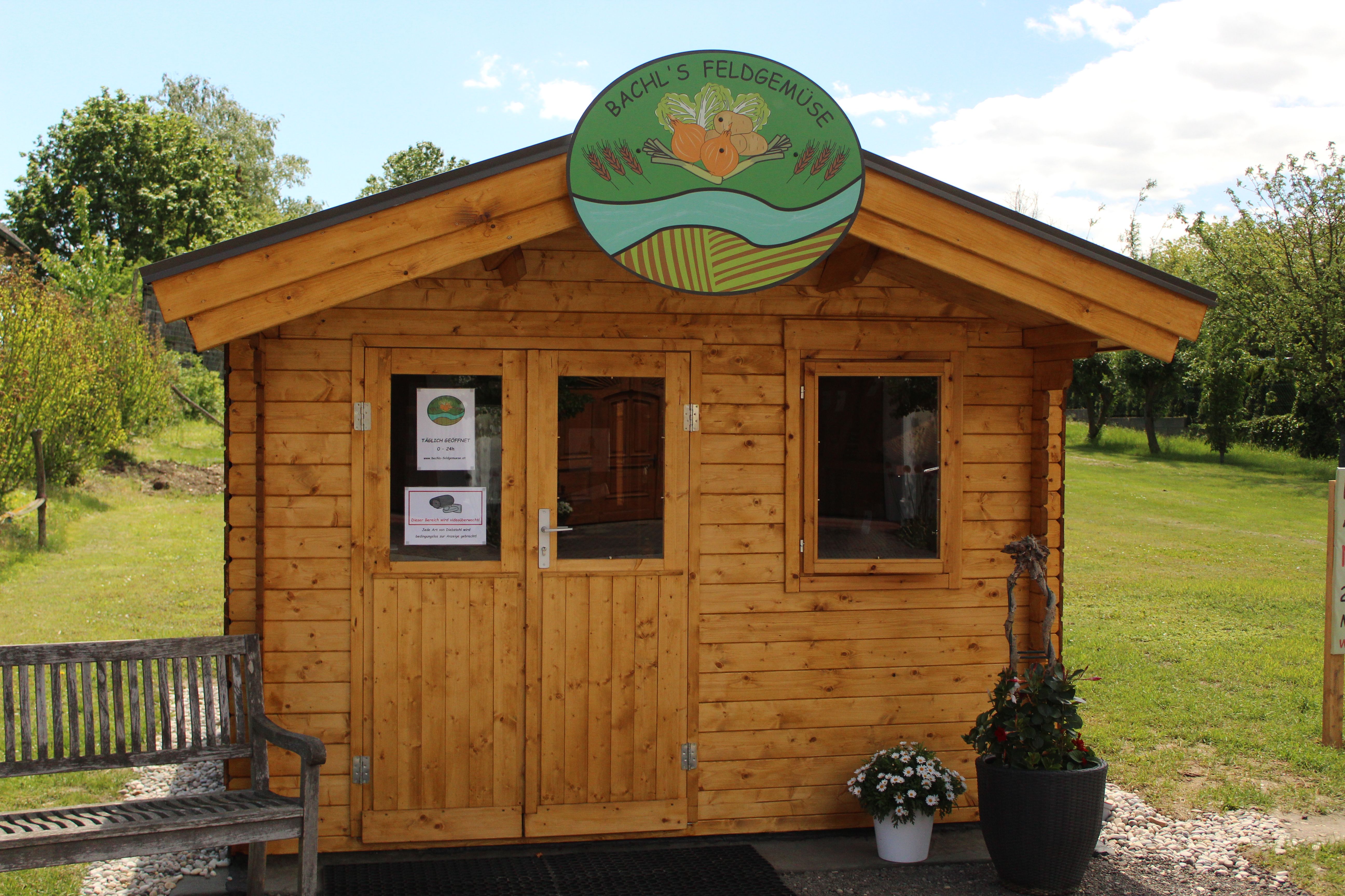 Wooden hut with sign 'Bachl's Feldgemüse' and flowers in front of it.