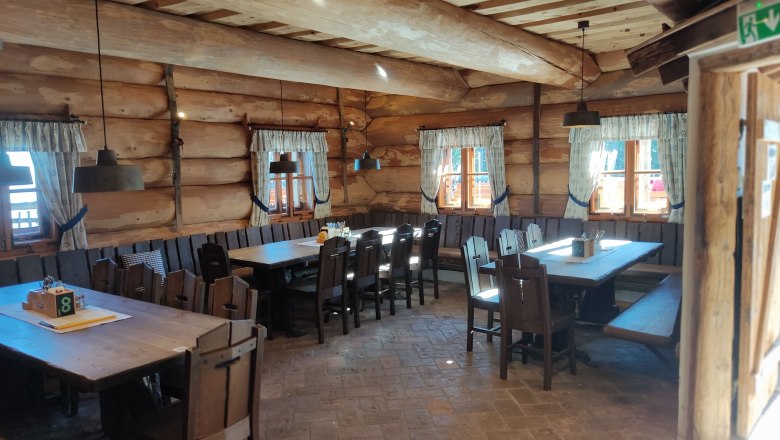 Interior view of a rustic wooden hut with wooden tables and chairs, decorated with curtains at the windows.