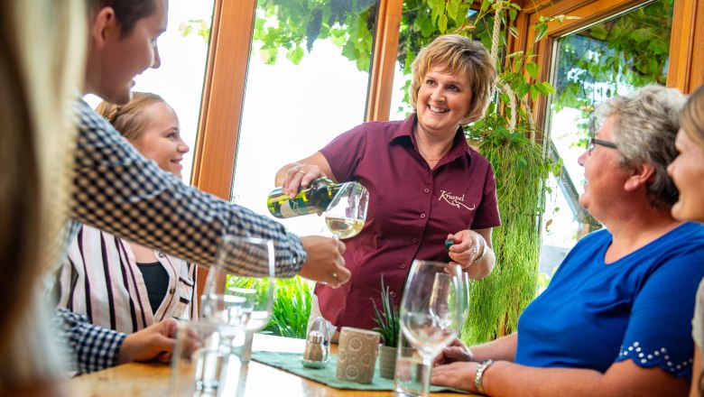 A woman pours wine for a group of people in a cozy room with plants.