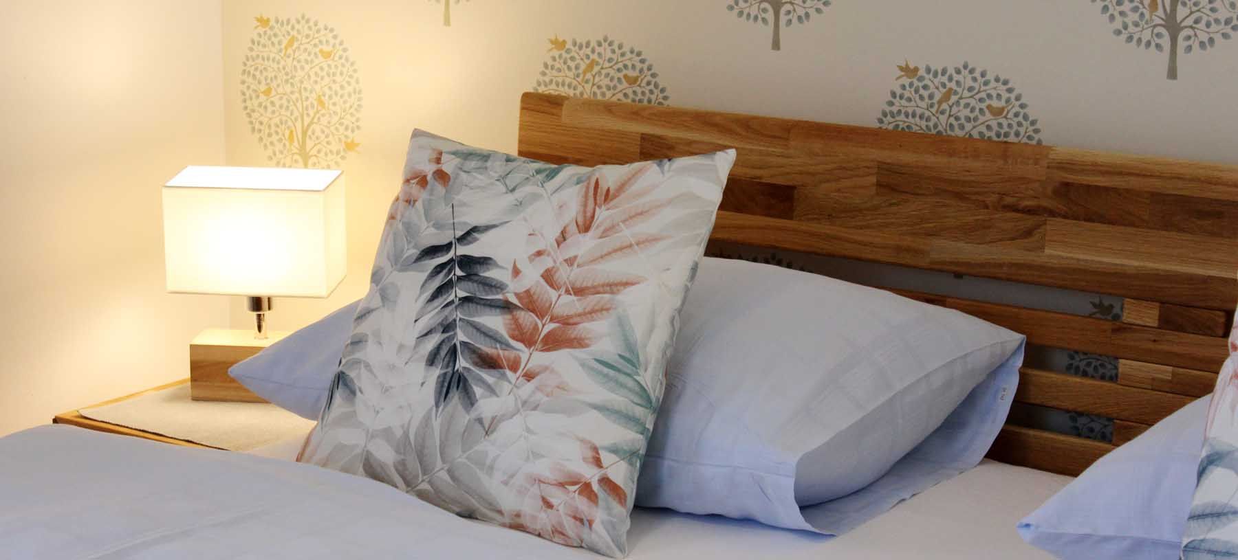 Close-up of a bed with wooden headboard, decorative cushions and bedside lamp.