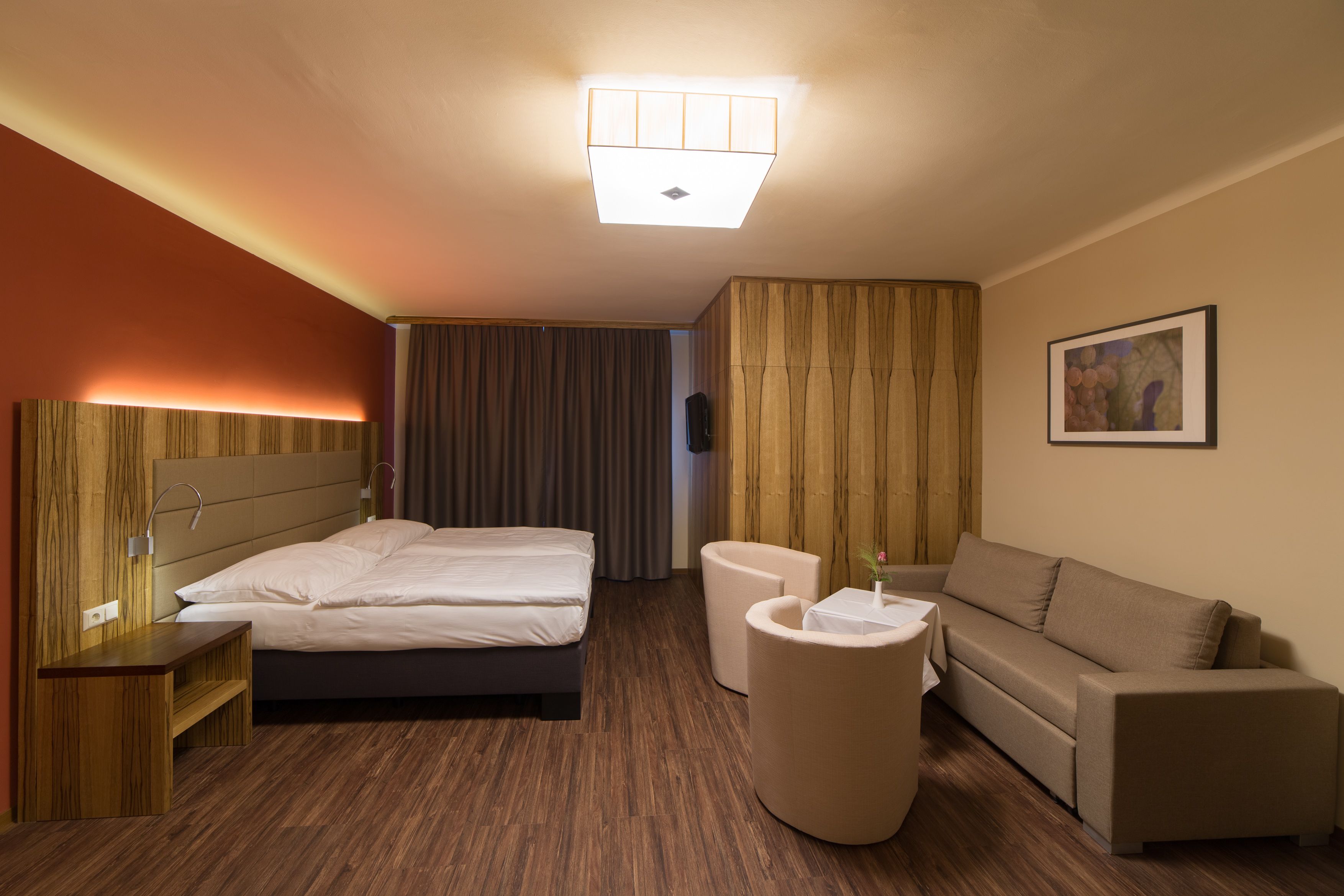 Modern furnished hotel room with double bed, sofa, two armchairs and a small table. Wooden floor and warm lighting.