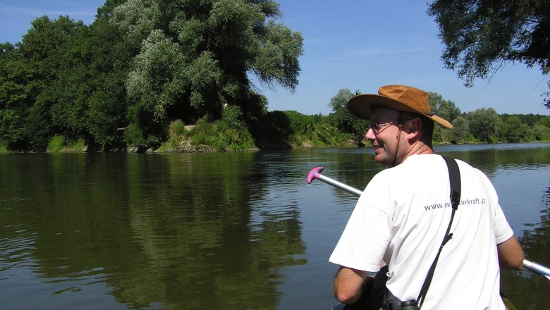 A man in a hat paddles in a canoe on a river surrounded by green trees.