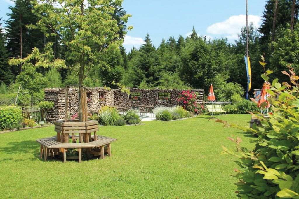 A green garden with wooden benches around a tree, surrounded by woods and flowers.
