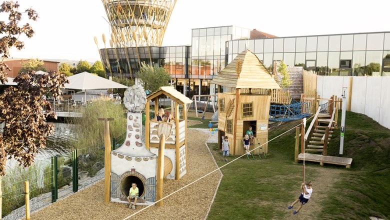 A modern playground with wooden houses and a rope slide in front of a building with a glass fa&ccedil;ade and a tower with wheat ornaments.