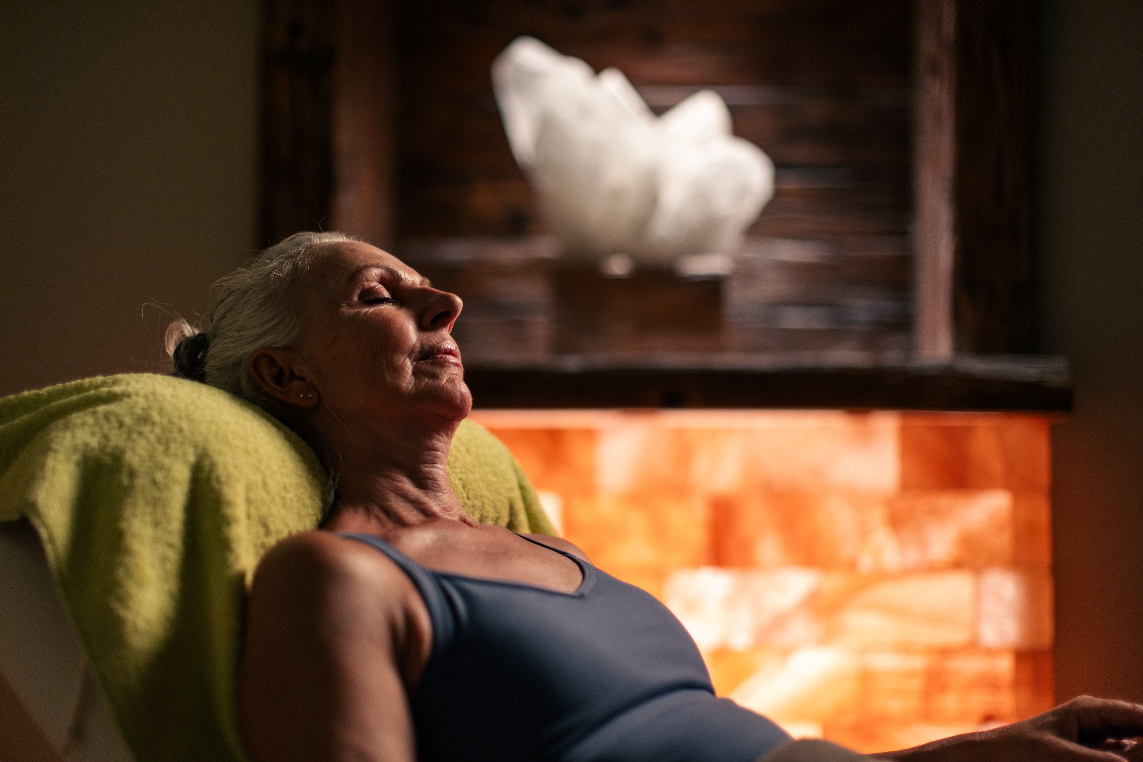 An elderly woman is relaxing at a spa, surrounded by warm light and a salt rock backdrop.