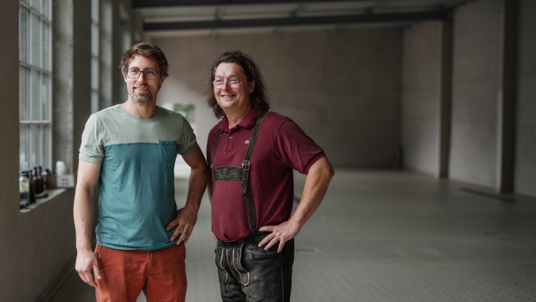 Two men, the founders of Raxbräu, stand in an empty room, one wearing a T-shirt and the other lederhosen.