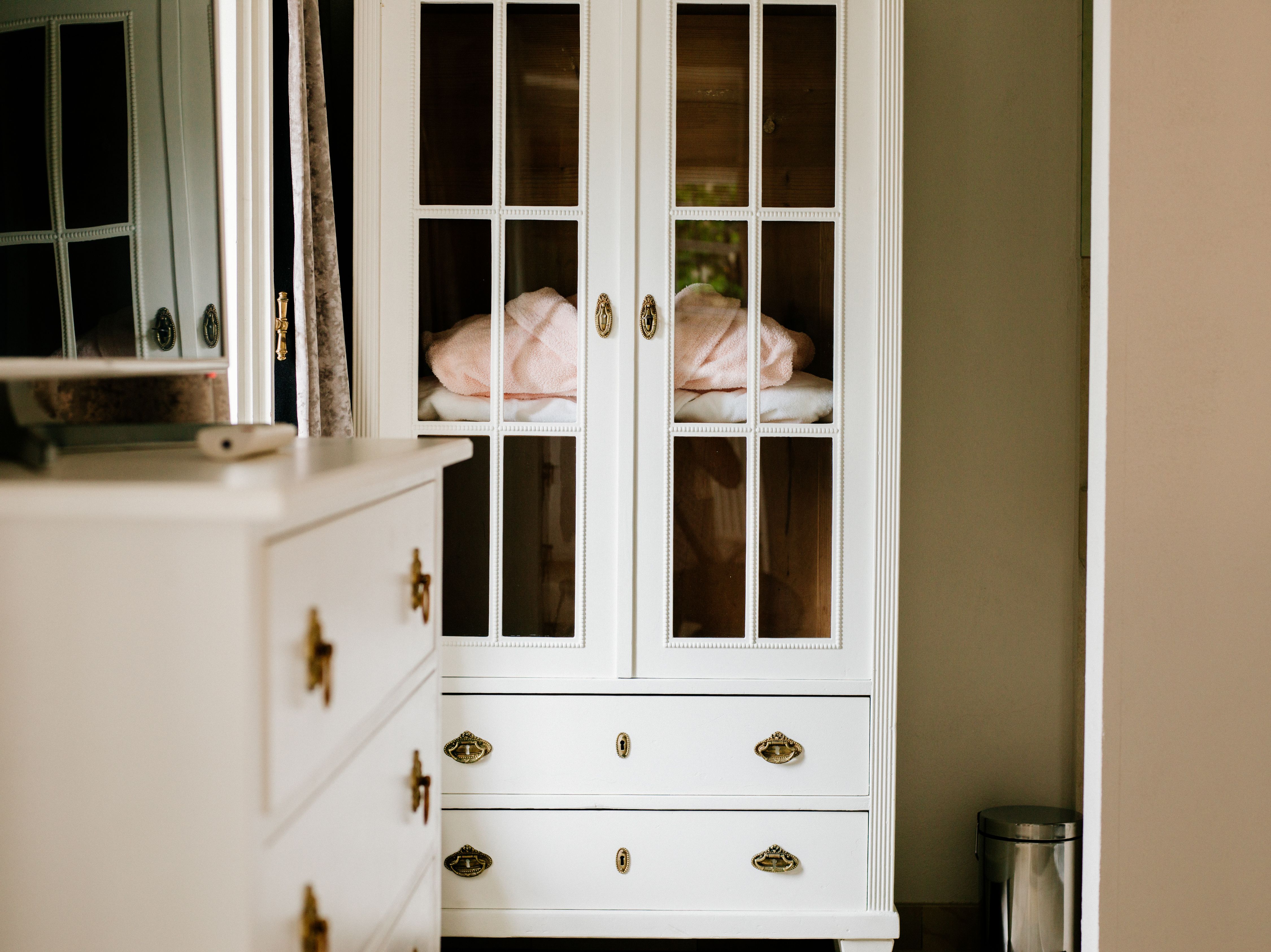 White cupboard with glass doors and drawers, next to it a chest of drawers and a waste garbage can.