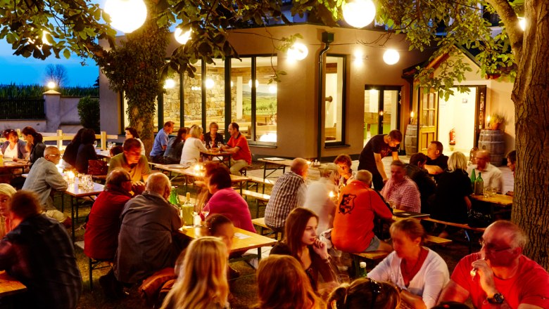 People sit at outdoor tables in the evening, surrounded by trees and lights.