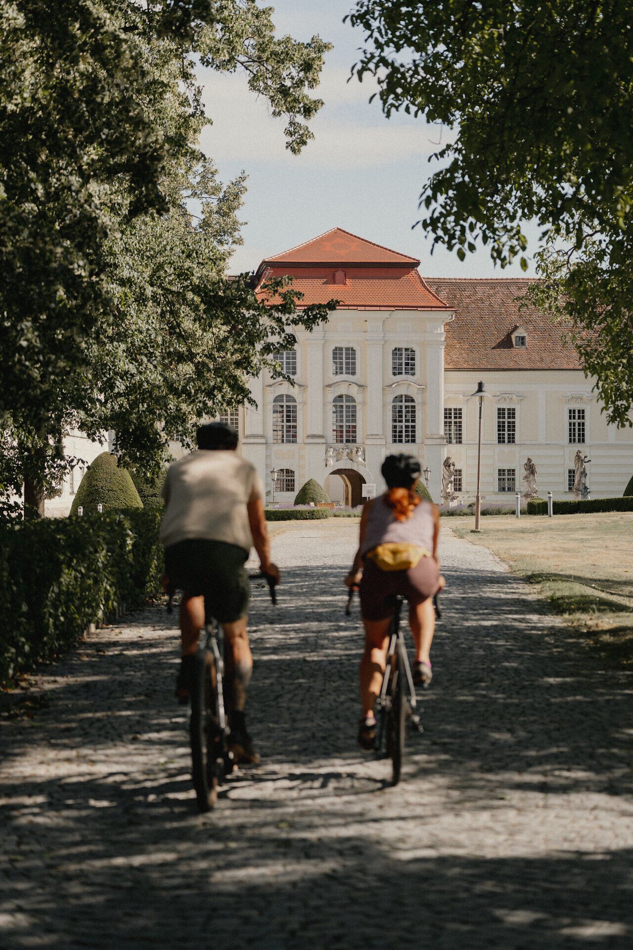 Two cyclists ride along a cobbled path towards a historic building surrounded by trees.