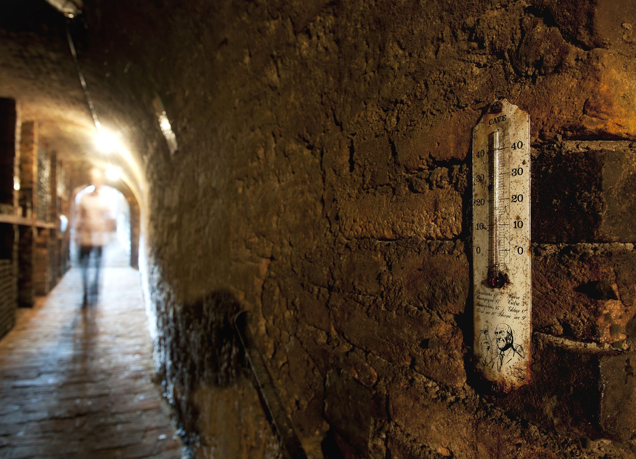 An old wine cellar with a thermometer on the wall and a blurred person in the background.