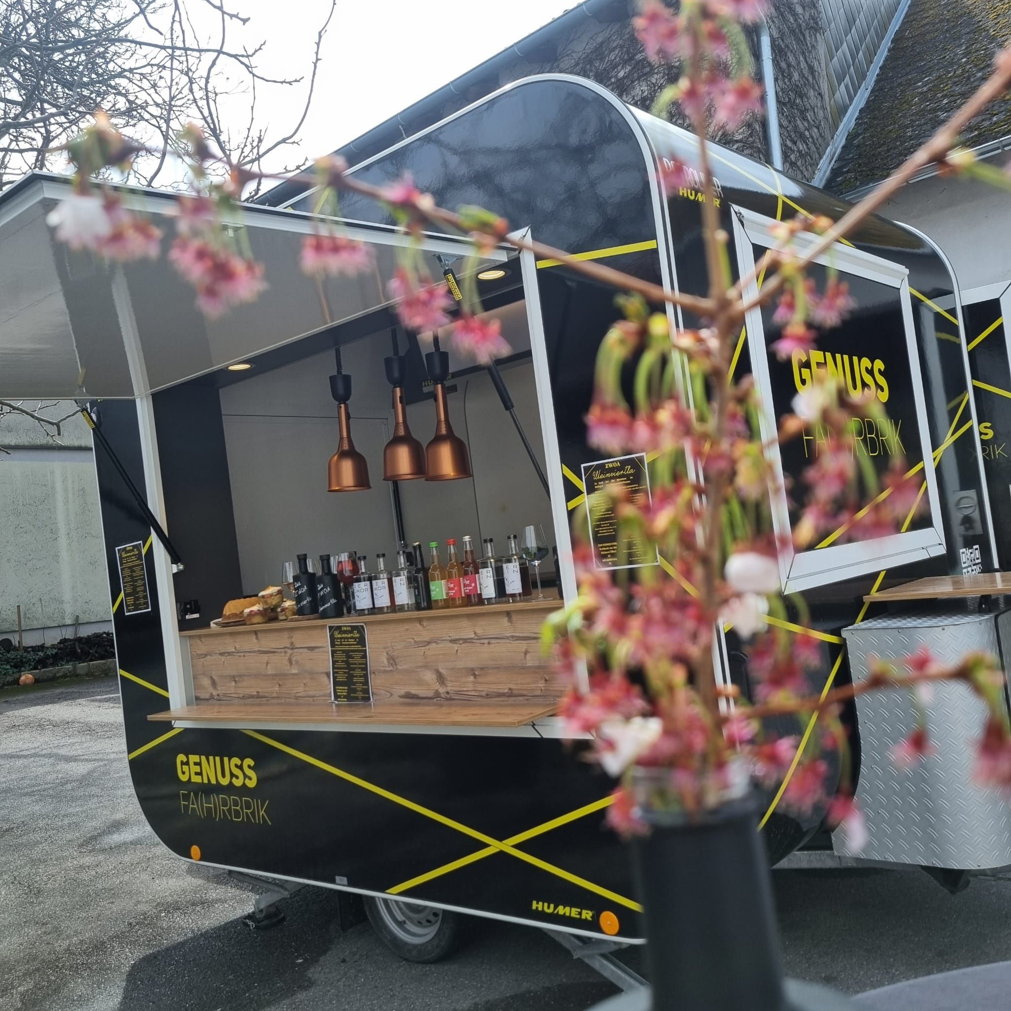 A black food truck with the inscription 'Genuss Fahrbrik' is parked outside. Blurred flowers can be seen in the foreground.