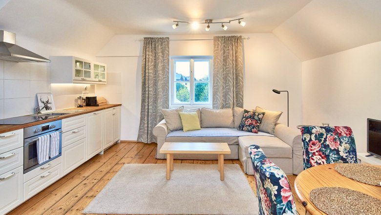 Cozy living room with open kitchen, wooden floor, beige couch, floral patterned chairs and a window with curtains.