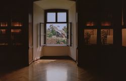 Blick aus einem historischen Raum im Schloss Grafenegg durch ein geöffnetes Fenster auf den modernen Wolkenturm, mit Bücherregalen und beleuchteten Vitrinen im Vordergrund.