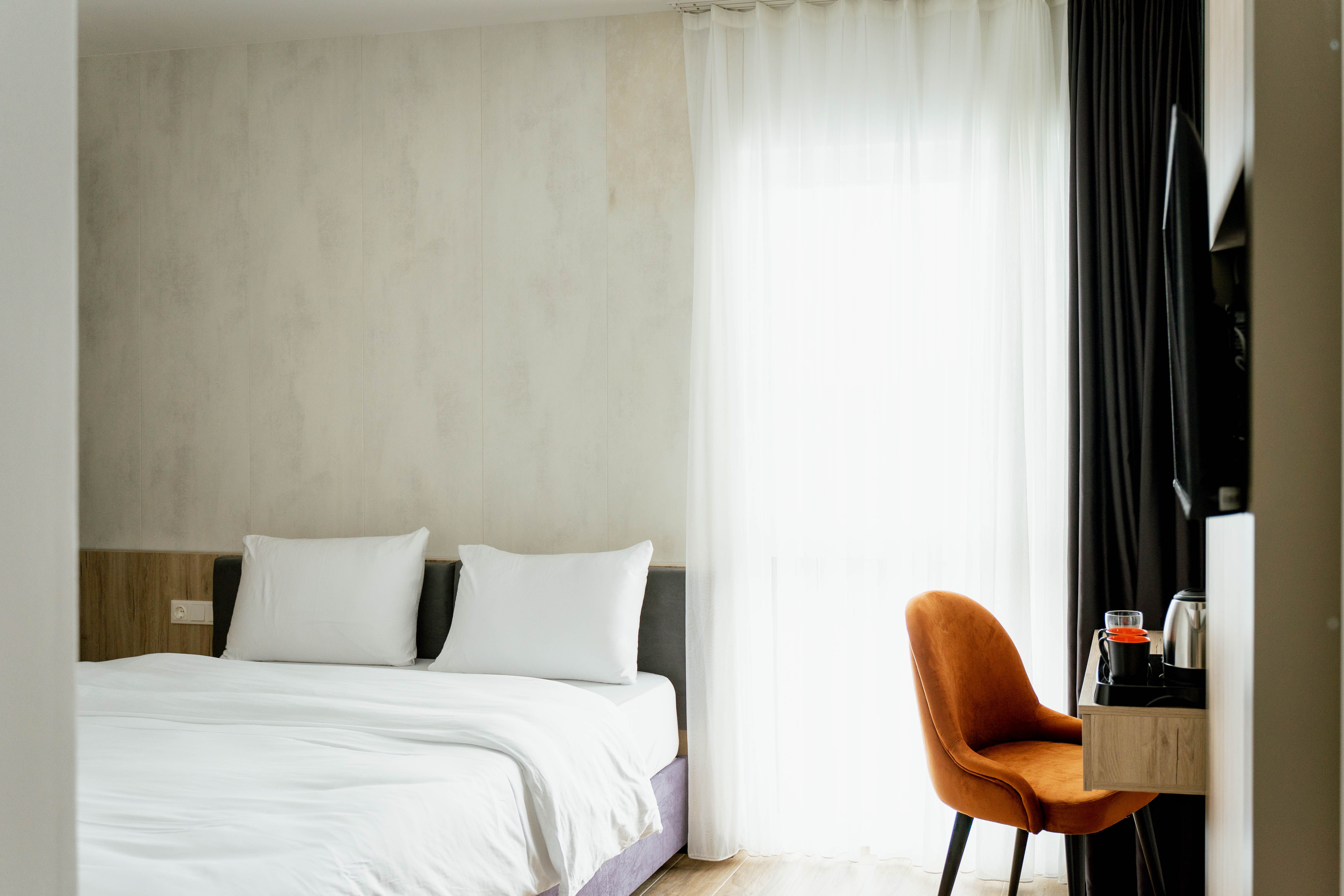 Modern hotel room with double bed, white sheets, wooden shelf and orange chair in front of a window with curtains.