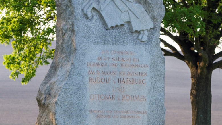 Memorial stone with knight relief and inscription, surrounded by trees and meadow.