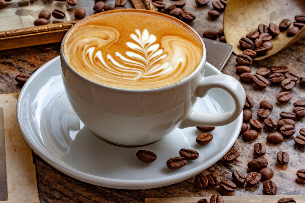 A cup of cappuccino with latte art on a table, surrounded by coffee beans.