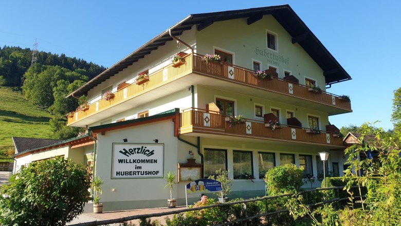 A traditional inn called Hubertushof with flowers on the balconies, surrounded by green countryside and blue skies.