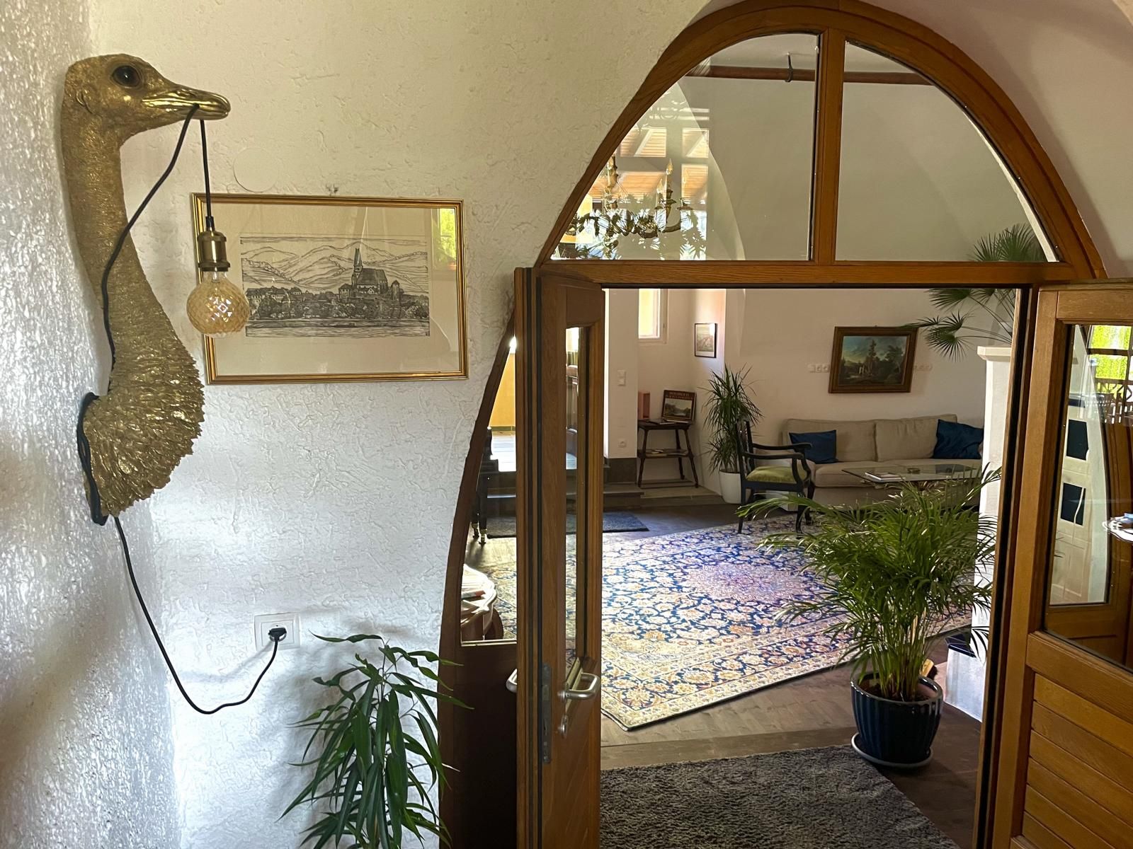 Interior view of a living room with plants, a sofa and a decorative wall object in the shape of a bird with a lamp.