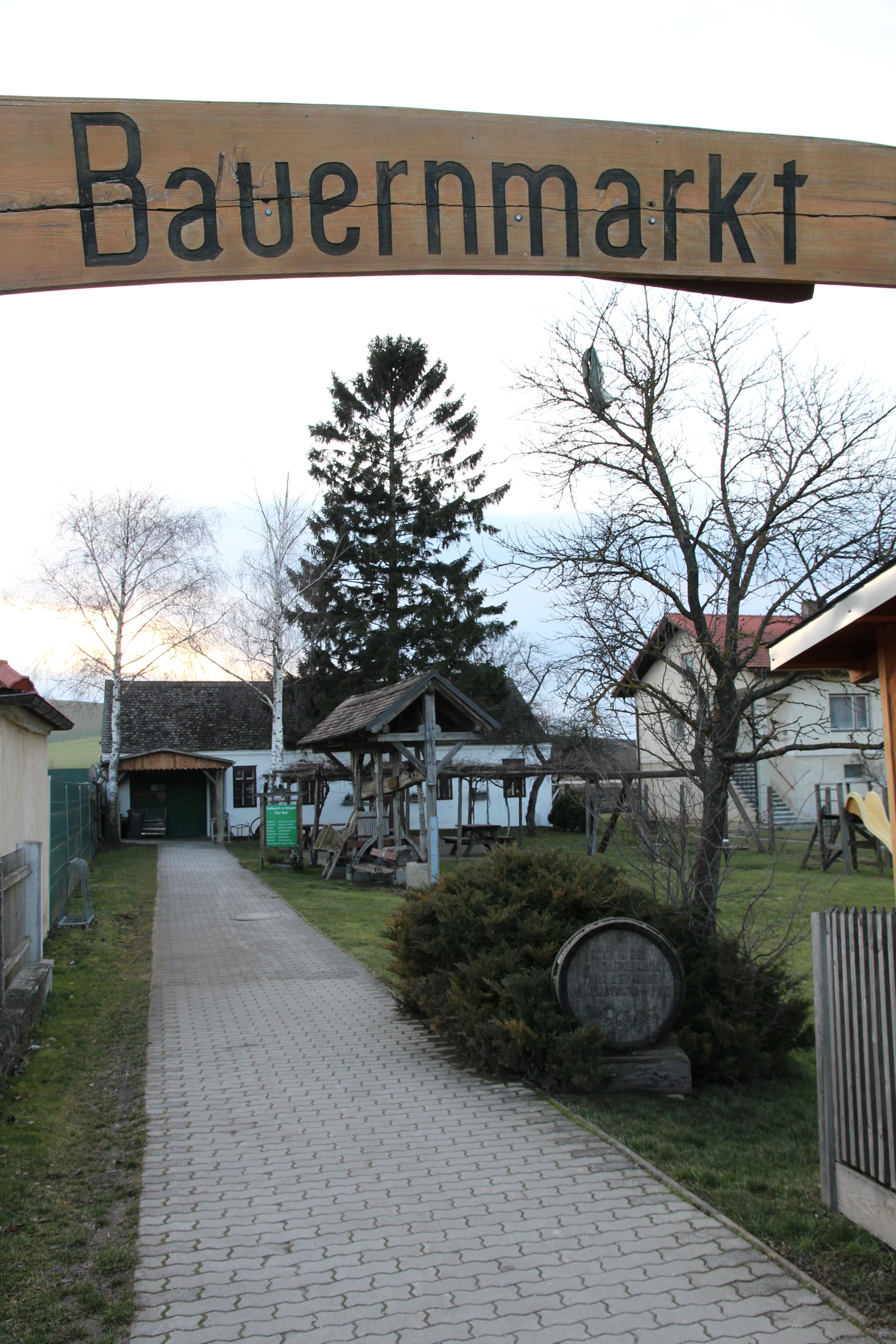 Entrance to a farmers' market with a wooden sign and a paved path surrounded by trees and buildings.